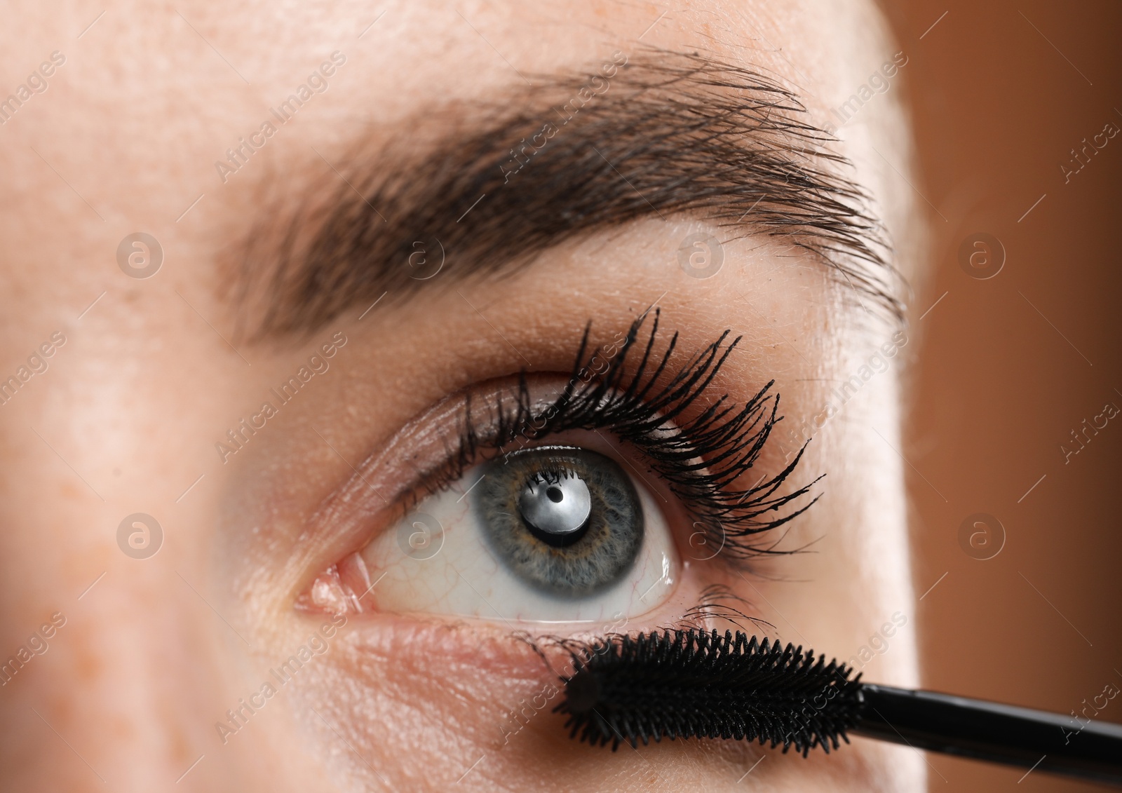 Young woman applying mascara onto her eyelashes on brown background, closeup Photo of Young woman applying mascara onto her eyelashes on brown background, closeup