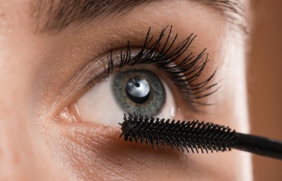 Photo of Young woman applying mascara onto her eyelashes on brown background, closeup