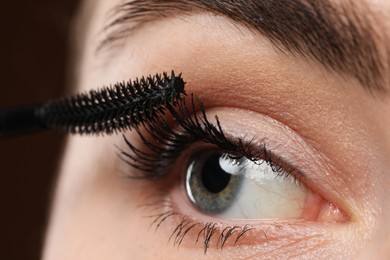 Photo of Young woman applying mascara onto her eyelashes on brown background, closeup