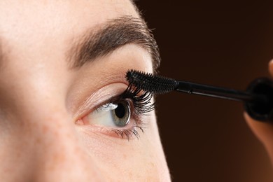 Photo of Young woman applying mascara onto her eyelashes on brown background, closeup