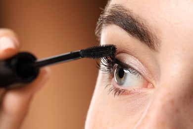 Photo of Young woman applying mascara onto her eyelashes on brown background, closeup