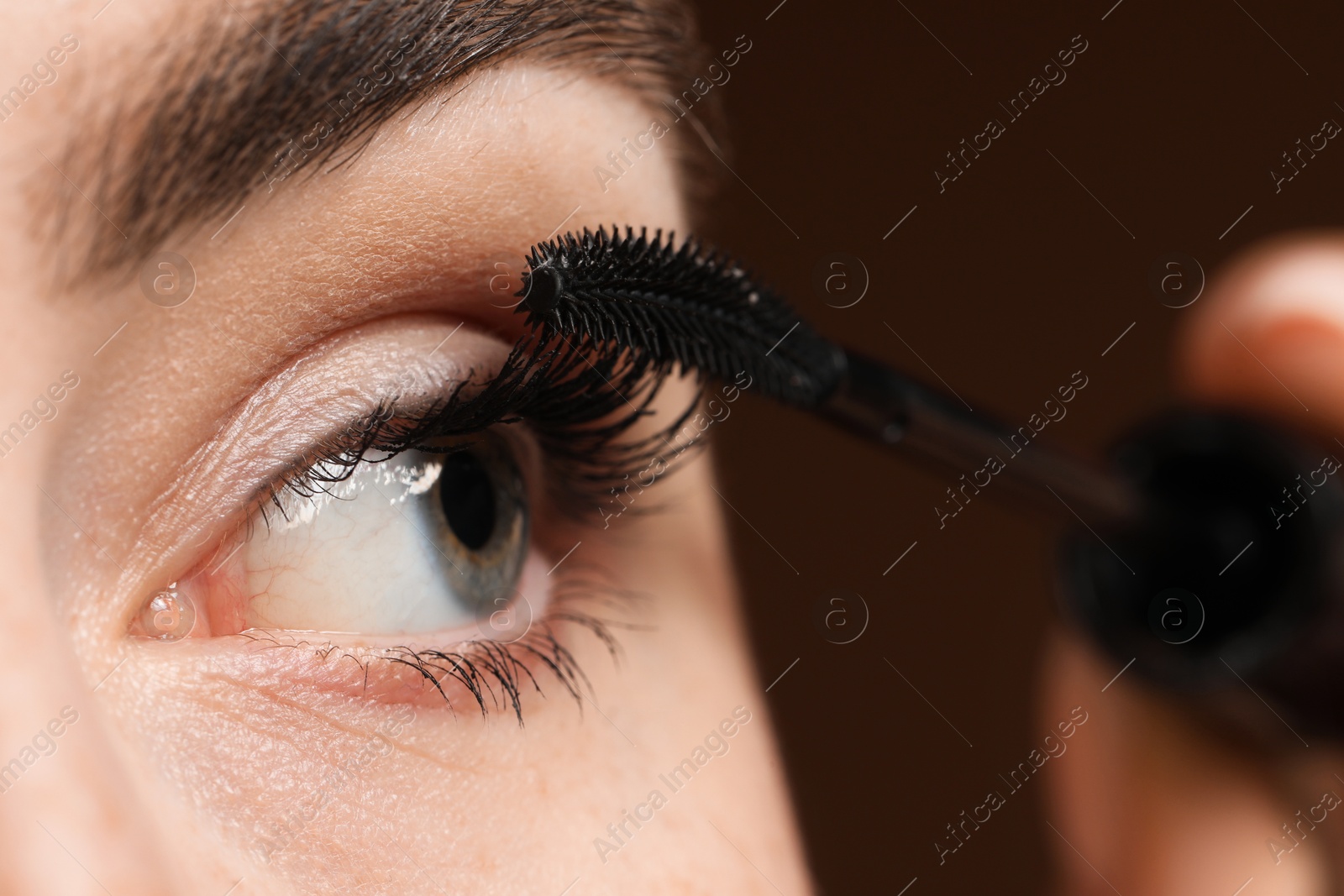 Young woman applying mascara onto her eyelashes on brown background, closeup Photo of Young woman applying mascara onto her eyelashes on brown background, closeup