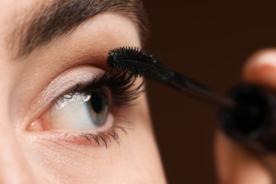 Photo of Young woman applying mascara onto her eyelashes on brown background, closeup