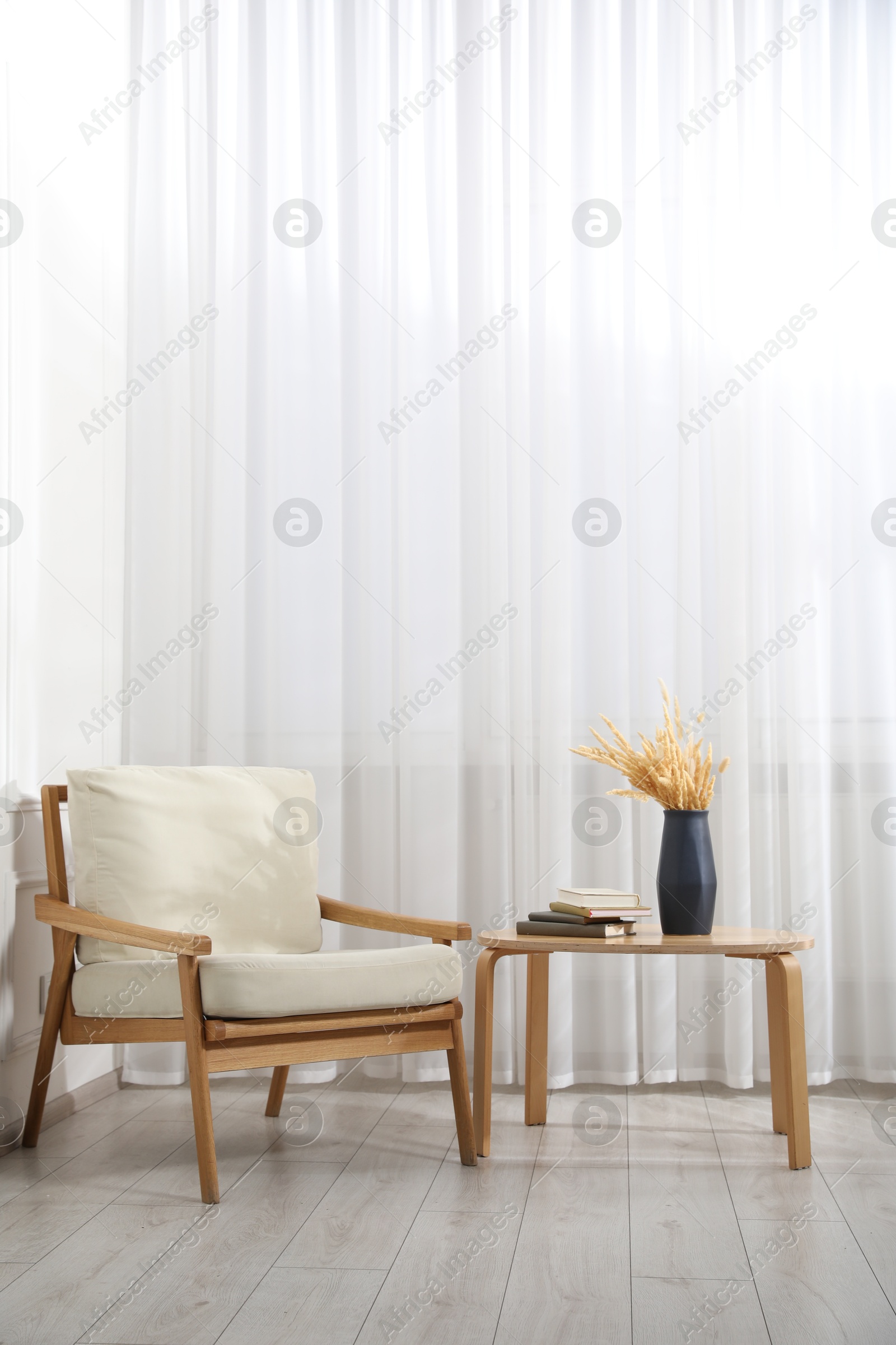 Comfortable beige armchair and table with vase and books near window indoors, space for text Photo of Comfortable beige armchair and table with vase and books near window indoors, space for text