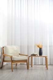 Photo of Comfortable beige armchair and table with vase and books near window indoors, space for text