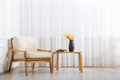 Photo of Comfortable beige armchair and table with vase and books near window indoors
