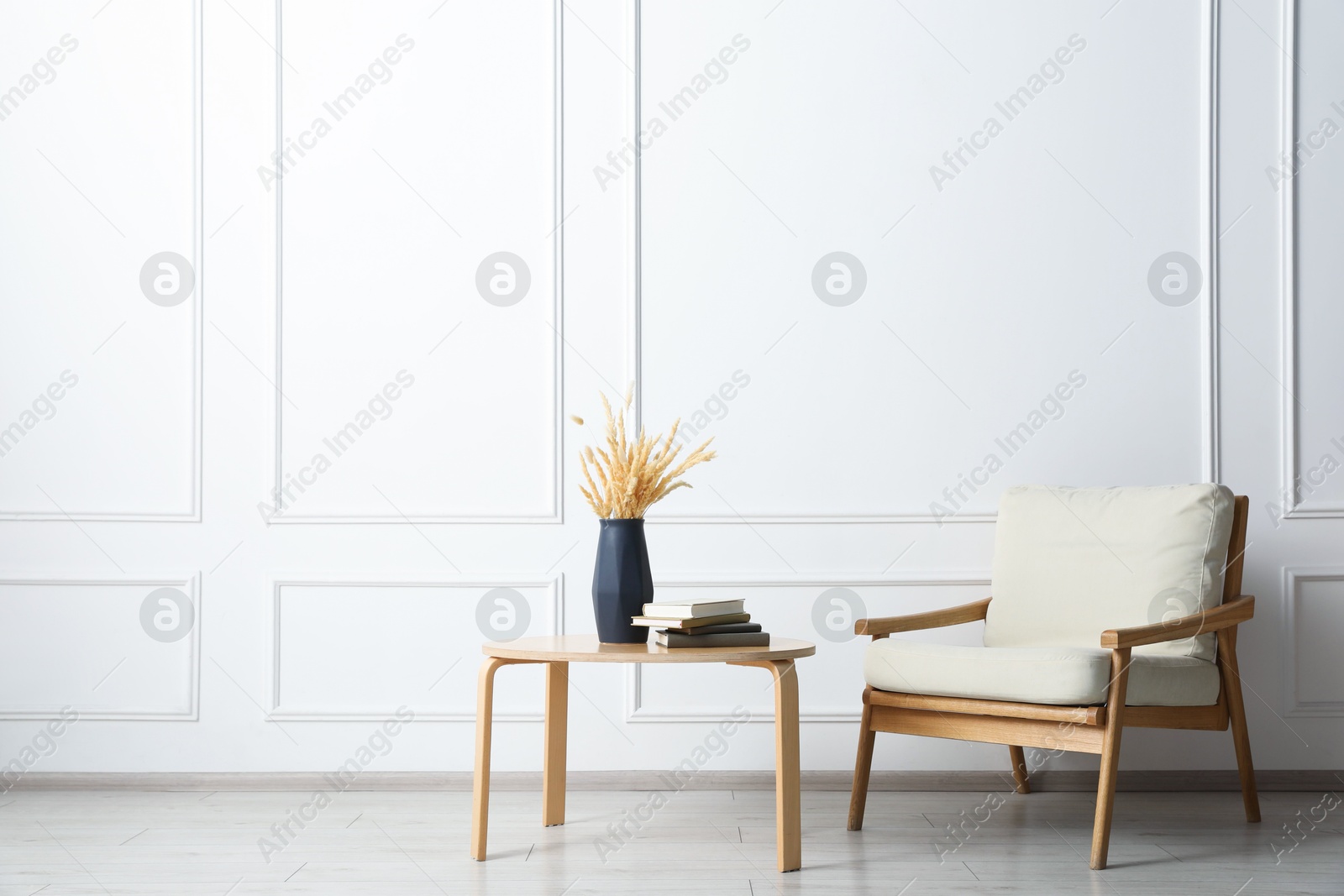 Comfortable beige armchair and table with vase and books near white wall indoors, space for text Photo of Comfortable beige armchair and table with vase and books near white wall indoors, space for text