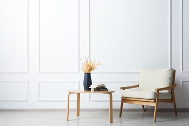 Photo of Comfortable beige armchair and table with vase and books near white wall indoors, space for text