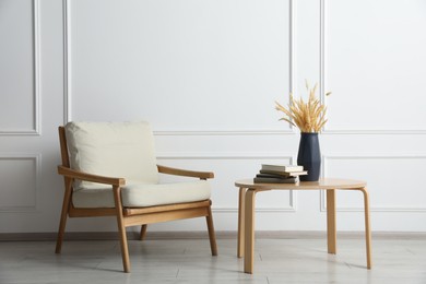 Photo of Comfortable beige armchair and table with vase and books near white wall indoors