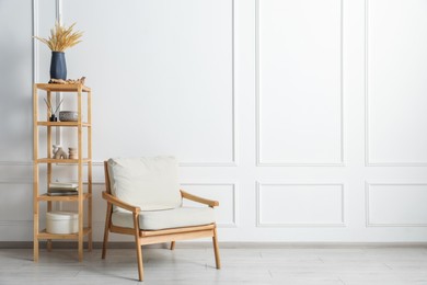 Photo of Comfortable beige armchair and wooden shelving unit with decor near white wall indoors, space for text