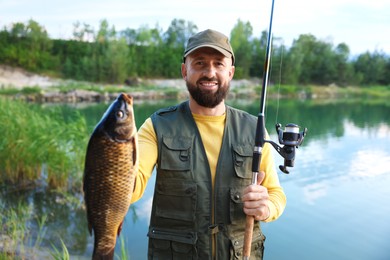 Photo of Smiling fisherman holding rod and caught fish near lake at summer