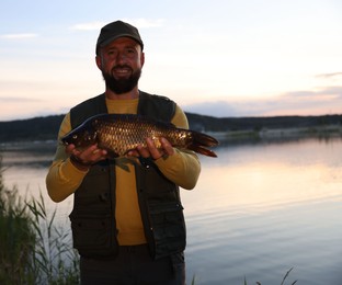 Photo of Smiling fisherman holding caught fish near lake at sunset