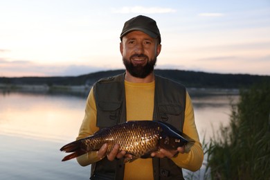 Photo of Smiling fisherman holding caught fish near lake at sunset
