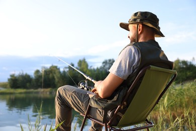 Photo of Fisherman with rod sitting on chair and fishing near lake at summer