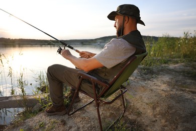 Photo of Fisherman with rod sitting on chair and fishing near lake at summer