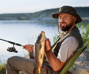 Photo of Smiling fisherman holding rod and caught fish near lake at summer