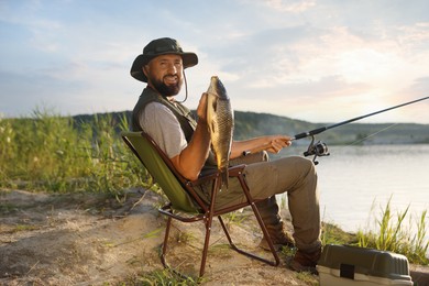 Photo of Smiling fisherman holding rod and caught fish near lake at summer