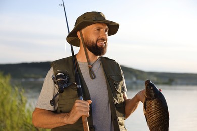 Photo of Smiling fisherman holding rod and caught fish near lake at summer