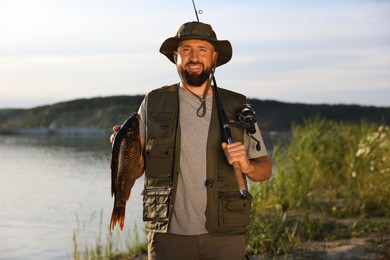Photo of Smiling fisherman holding rod and caught fish near lake at summer
