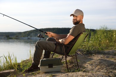 Photo of Fisherman with rod sitting on chair and fishing near lake at summer