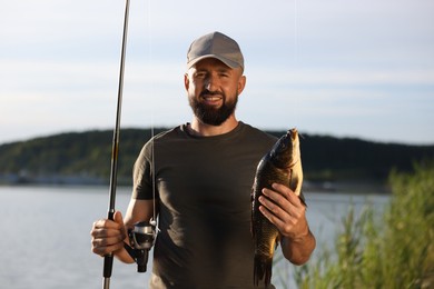 Photo of Smiling fisherman holding rod and caught fish near lake at summer