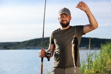 Photo of Smiling fisherman holding rod and caught fish near lake at summer