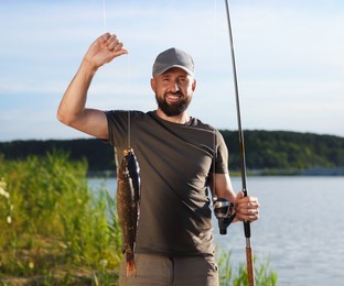 Photo of Smiling fisherman holding rod and caught fish near lake at summer