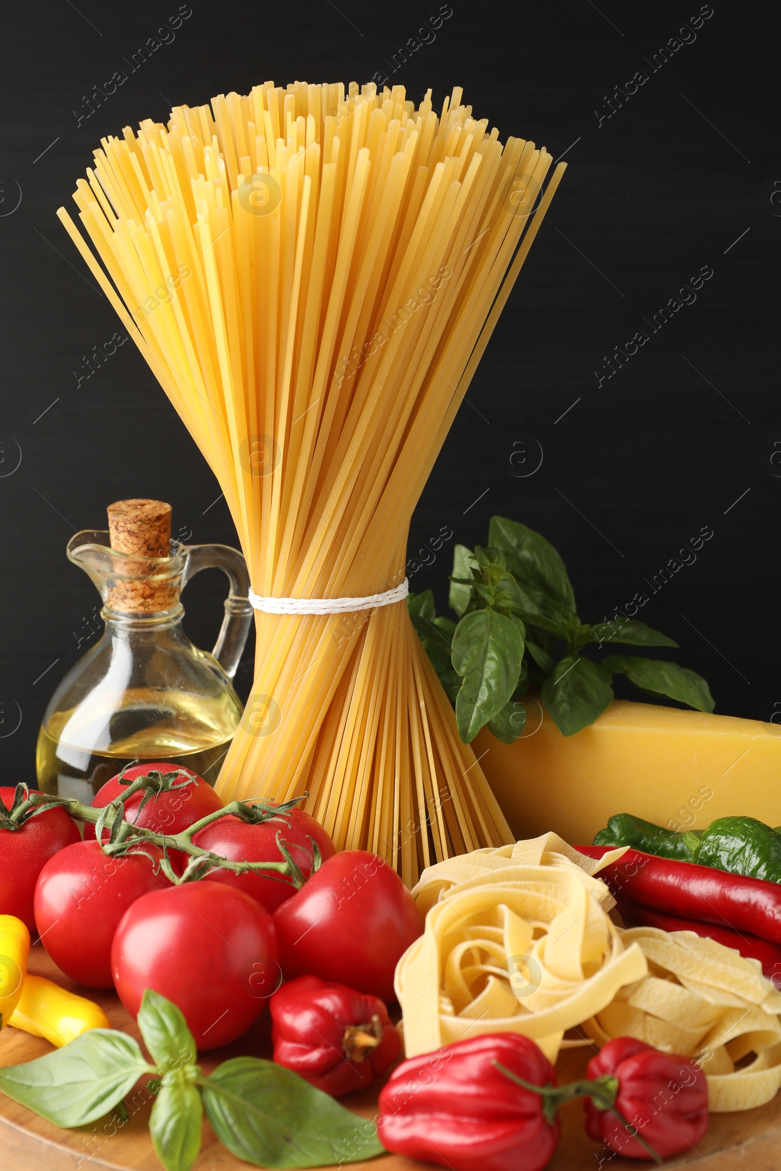 Photo of Different types of raw pasta and products on wooden board against black background, closeup