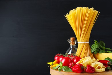Photo of Different types of raw pasta and products on wooden board against black background, closeup. Space for text
