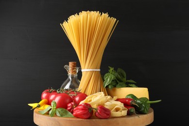 Photo of Different types of raw pasta and products on wooden board against black background, closeup