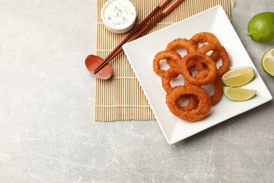 Photo of Fried squid rings, lime, sauce and chopsticks on grey table, flat lay. Space for text