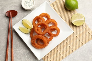 Photo of Fried squid rings, lime, sauce and chopsticks on grey table, flat lay