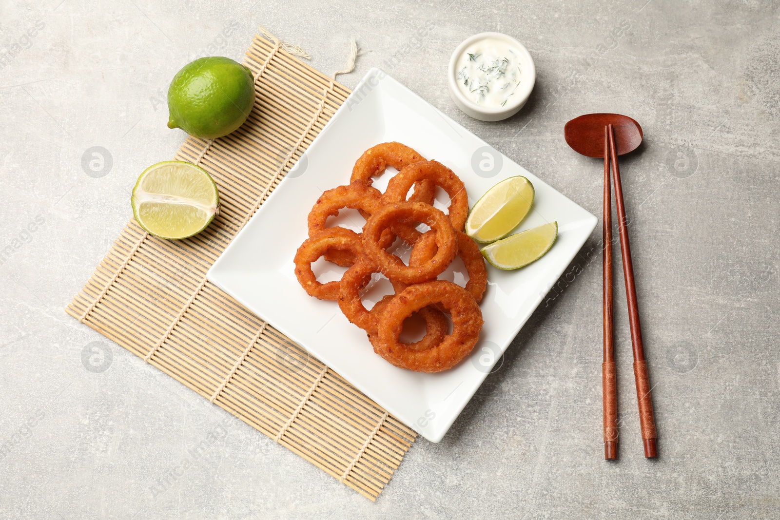 Photo of Fried squid rings, lime, sauce and chopsticks on grey table, flat lay