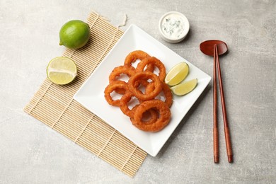 Photo of Fried squid rings, lime, sauce and chopsticks on grey table, flat lay