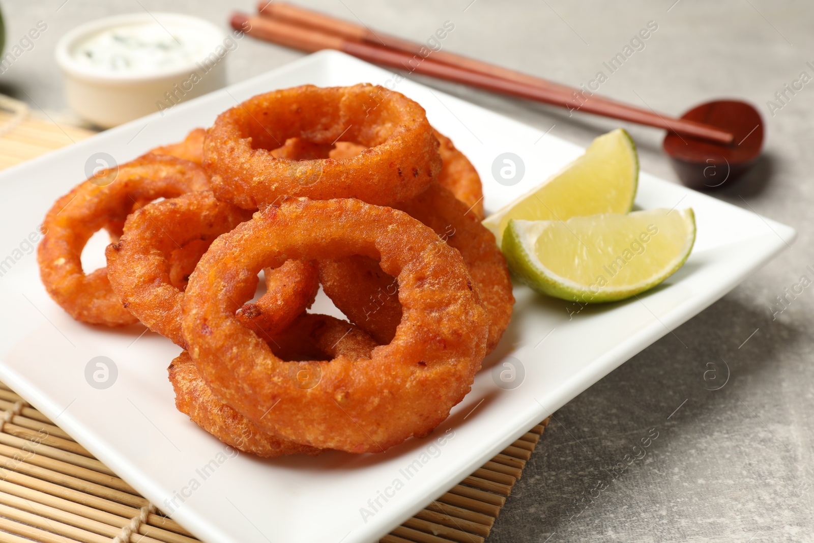 Photo of Fried squid rings, lime, sauce and chopsticks on grey table, closeup