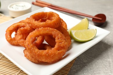 Photo of Fried squid rings, lime, sauce and chopsticks on grey table, closeup