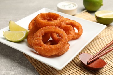 Photo of Fried squid rings, lime, sauce and chopsticks on grey table, closeup