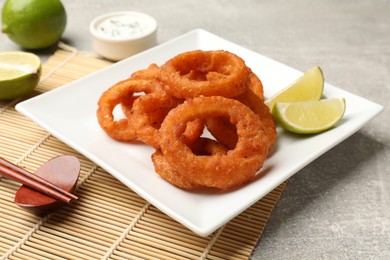 Photo of Fried squid rings, lime, sauce and chopsticks on grey table, closeup