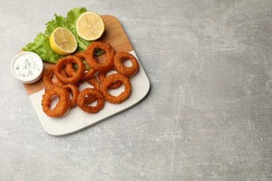 Photo of Fried squid rings, lemon, lettuce and sauce on grey table, top view. Space for text