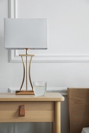 Photo of Bedside table with lamp and glass of water near bed indoors