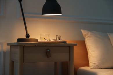 Photo of Bedside table with lamp, notebook, glasses and glass of water near bed indoors