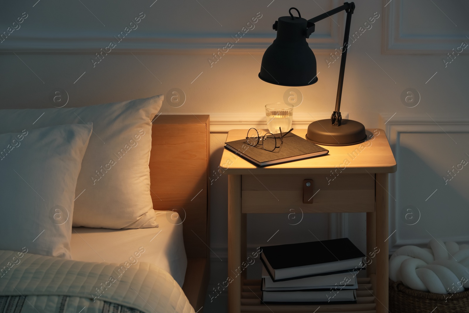 Photo of Bedside table with lamp, notebook, glasses and glass of water near bed indoors