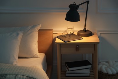 Photo of Bedside table with lamp, notebook, glasses and glass of water near bed indoors
