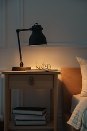 Photo of Bedside table with lamp, notebook, glasses and glass of water near bed indoors