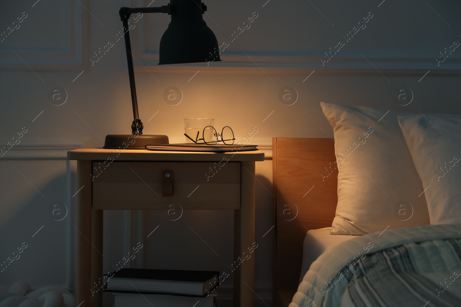 Photo of Bedside table with lamp, notebook, glasses and glass of water near bed indoors