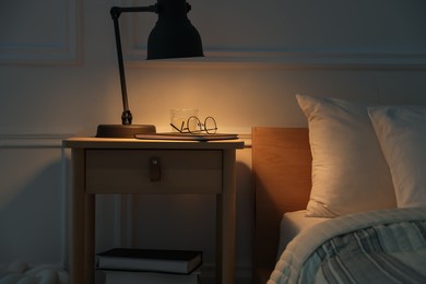 Photo of Bedside table with lamp, notebook, glasses and glass of water near bed indoors