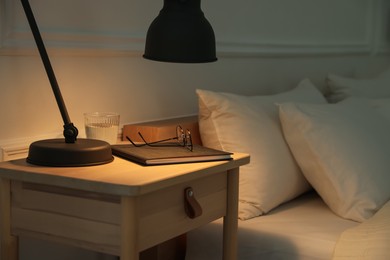 Photo of Bedside table with lamp, notebook, glasses and glass of water near bed indoors