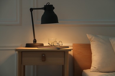 Photo of Bedside table with lamp, notebook, glasses and glass of water near bed indoors
