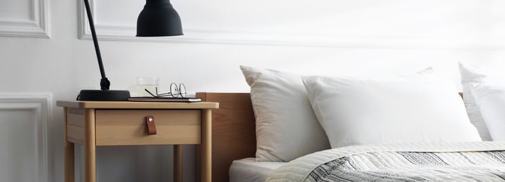 Photo of Bedside table with lamp, notebook, glasses and glass of water near bed indoors