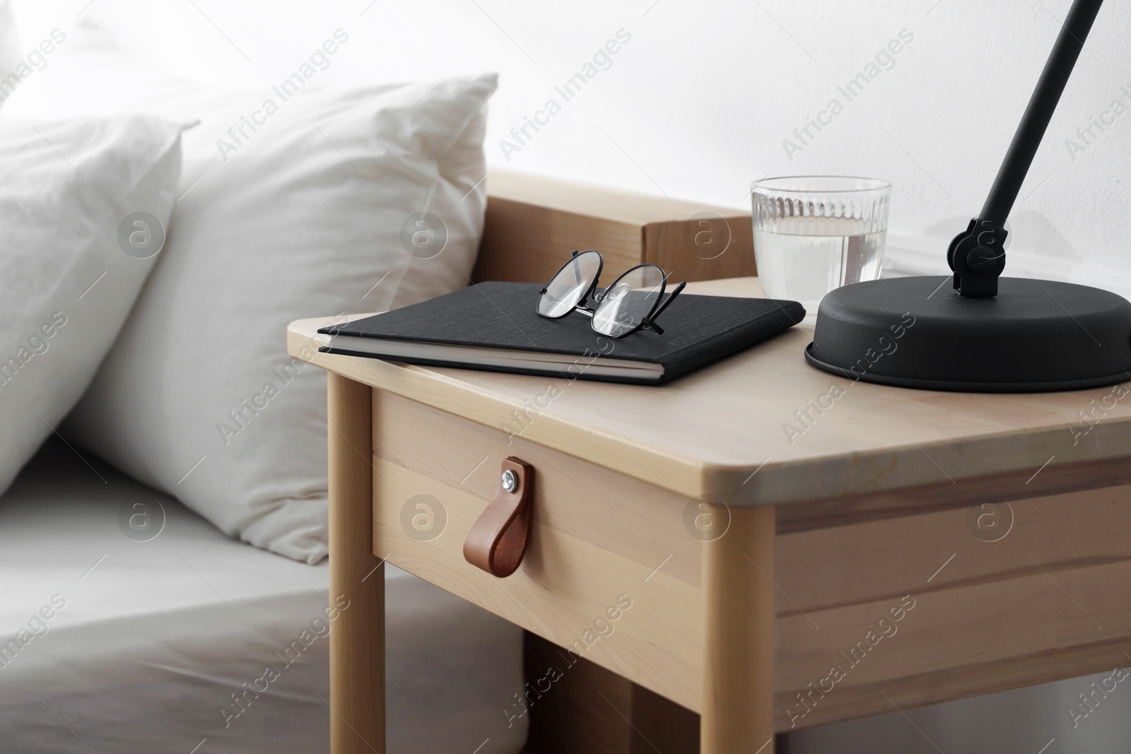 Photo of Bedside table with lamp, notebook, glasses and glass of water near bed indoors, closeup